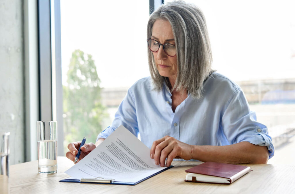 A senior woman in a button-up shirt reviewing a document on her table.