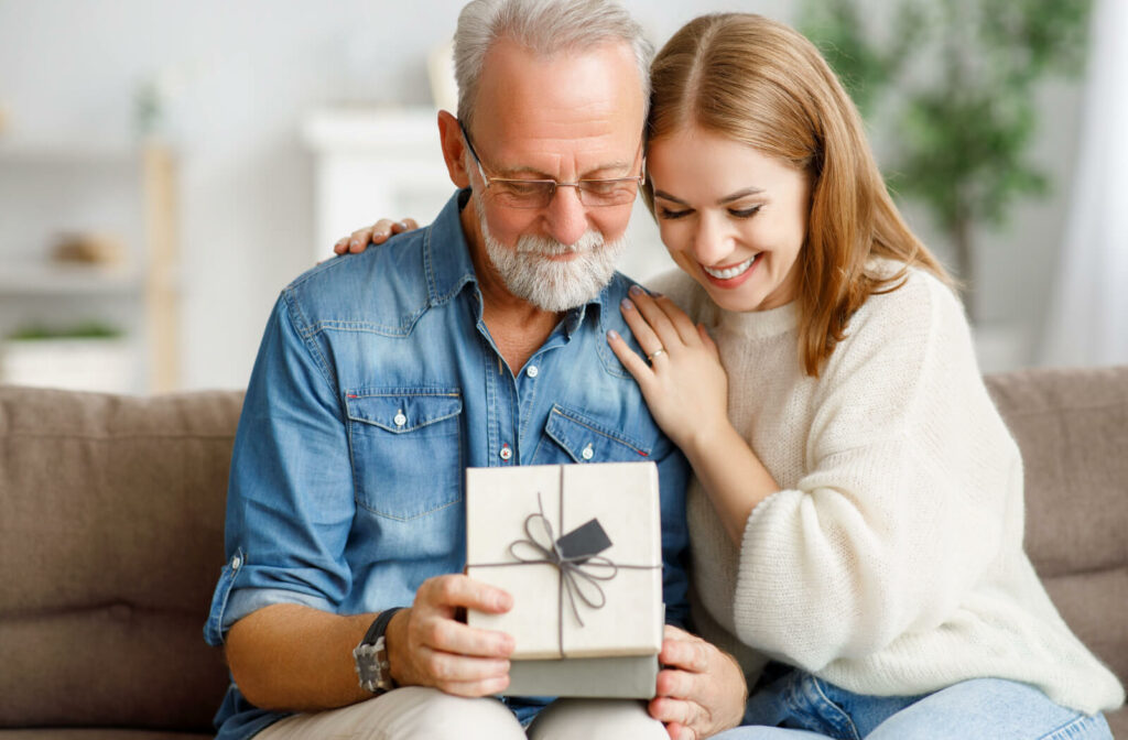 An adult child sitting beside a senior parent, smiling and hugging as the parent opens a white gift box.