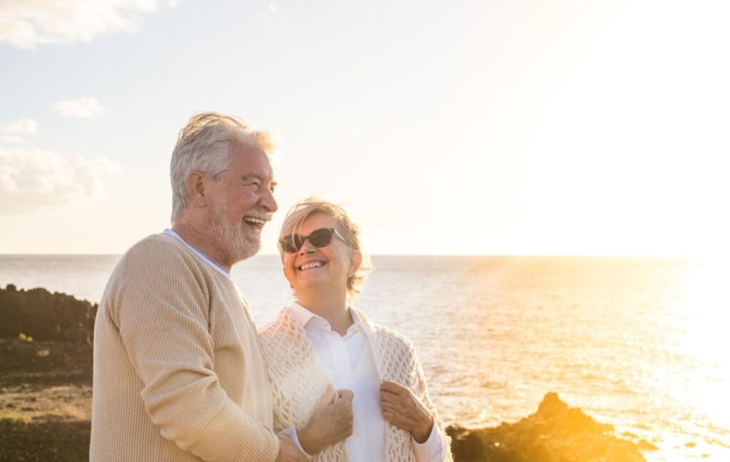 A senior couple laugh and smile as they walk down a picturesque New Jersey beach together during a golden sunset.